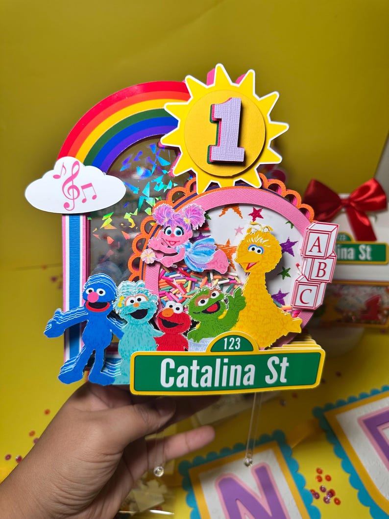 K&ouml;nnte beinhalten: Ein farbenfroher Tortenaufsatz zum Geburtstag mit Sesamstra&szlig;en-Figuren, einem Regenbogen und einer Sonne mit der Zahl 1. Der Aufsatz enth&auml;lt ein Stra&szlig;enschild mit der Aufschrift "Catalina St". Das Design ist lebendig mit Rosa, Gelb, Blau und Gr&uuml;n.