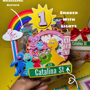 K&ouml;nnte beinhalten: Ein farbenfroher, mehrschichtiger Geburtstagskuchenaufsatz mit Sesamstra&szlig;en-Charakteren. Der Aufsatz enth&auml;lt einen Regenbogen, eine Sonne mit der Zahl 1 und ein Stra&szlig;enschild mit der Aufschrift "Catalina St". Das Design beinhaltet holografisches Acetat und einen Shaker mit Lichtern.