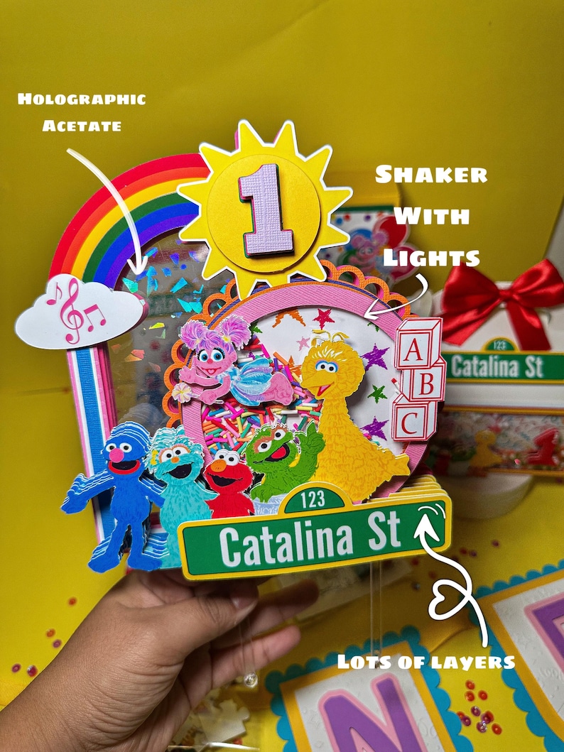 K&ouml;nnte beinhalten: Ein farbenfroher, mehrschichtiger Geburtstagskuchenaufsatz mit Sesamstra&szlig;en-Charakteren. Der Aufsatz enth&auml;lt einen Regenbogen, eine Sonne mit der Zahl 1 und ein Stra&szlig;enschild mit der Aufschrift "Catalina St". Das Design beinhaltet holografisches Acetat und einen Shaker mit Lichtern.