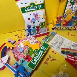 K&ouml;nnte beinhalten: Eine farbenfrohe Geburtstagsfeier mit Charakteren aus der Sesamstra&szlig;e. Enth&auml;lt eine Snackt&uuml;te mit dem Text "Catalina St", eine Eins und verschiedene Dekorationen. Die Szene spielt sich auf einer gelben Oberfl&auml;che mit Konfetti ab.