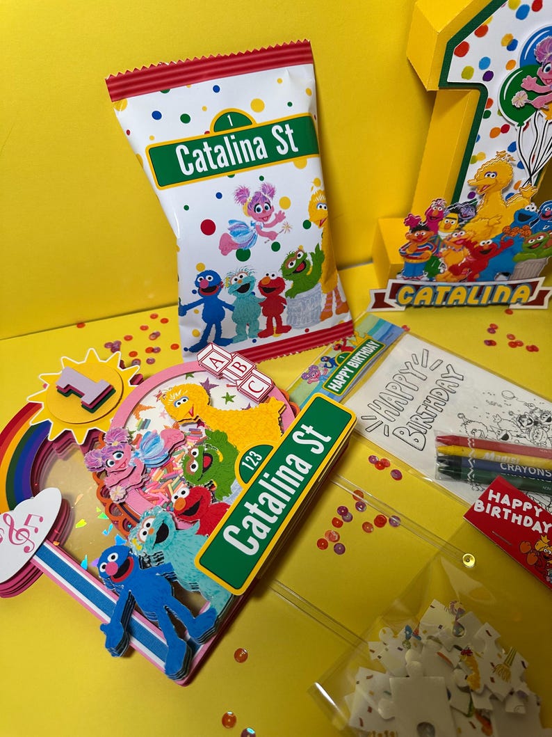 K&ouml;nnte beinhalten: Eine farbenfrohe Geburtstagsfeier mit Charakteren aus der Sesamstra&szlig;e. Enth&auml;lt eine Snackt&uuml;te mit dem Text "Catalina St", eine Eins und verschiedene Dekorationen. Die Szene spielt sich auf einer gelben Oberfl&auml;che mit Konfetti ab.