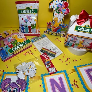 K&ouml;nnte beinhalten: Eine Sammlung von Partyzubeh&ouml;r zum Thema Sesamstra&szlig;e, darunter eine T&uuml;te mit Leckereien, eine Eins-Aussparung, eine Geschenkbox und ein Banner. Die Artikel zeigen Charaktere wie Elmo und Big Bird mit dem Text "Catalina St" und "Happy Birthday".