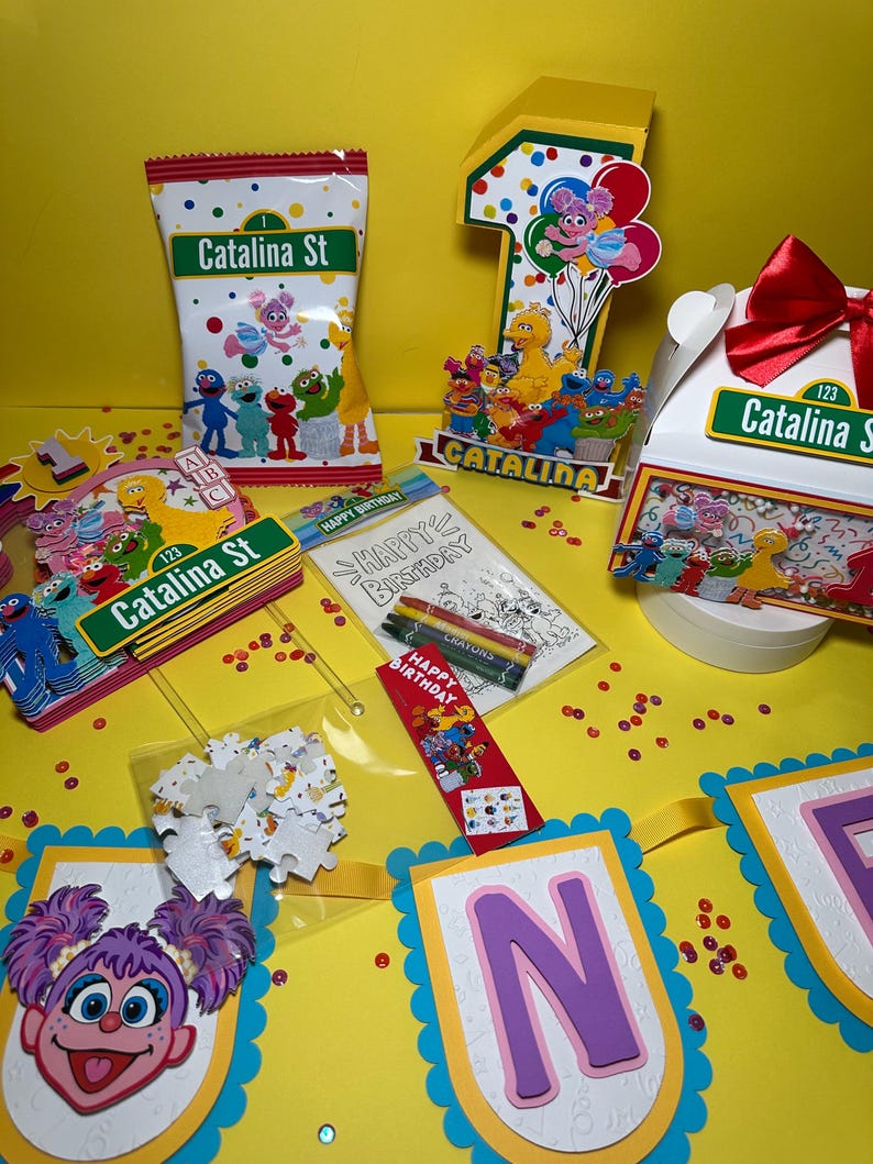K&ouml;nnte beinhalten: Eine Sammlung von Partyzubeh&ouml;r zum Thema Sesamstra&szlig;e, darunter eine T&uuml;te mit Leckereien, eine Eins-Aussparung, eine Geschenkbox und ein Banner. Die Artikel zeigen Charaktere wie Elmo und Big Bird mit dem Text "Catalina St" und "Happy Birthday".