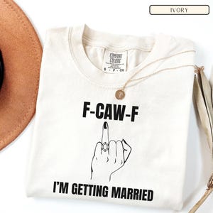 May include: Ivory-colored t-shirt with the text "F-CAW-F" above a graphic of a hand with a ring, giving the middle finger. Below the graphic is the text "I'M GETTING MARRIED". A gold necklace and a brown hat are also visible.