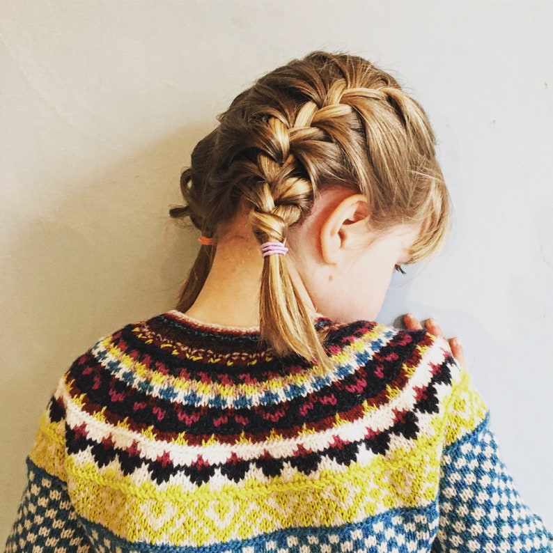 Puede incluir: Una persona joven con el pelo largo y rubio peinado en una trenza francesa, lleva un su&eacute;ter de punto colorido con un patr&oacute;n geom&eacute;trico.