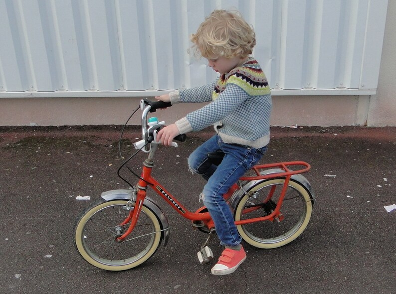 Puede incluir: Una persona joven montando una bicicleta roja con neum&aacute;ticos blancos y un asiento negro. La bicicleta tiene una campana plateada y un manillar cromado. La persona lleva un su&eacute;ter azul y blanco y pantalones vaqueros azules.