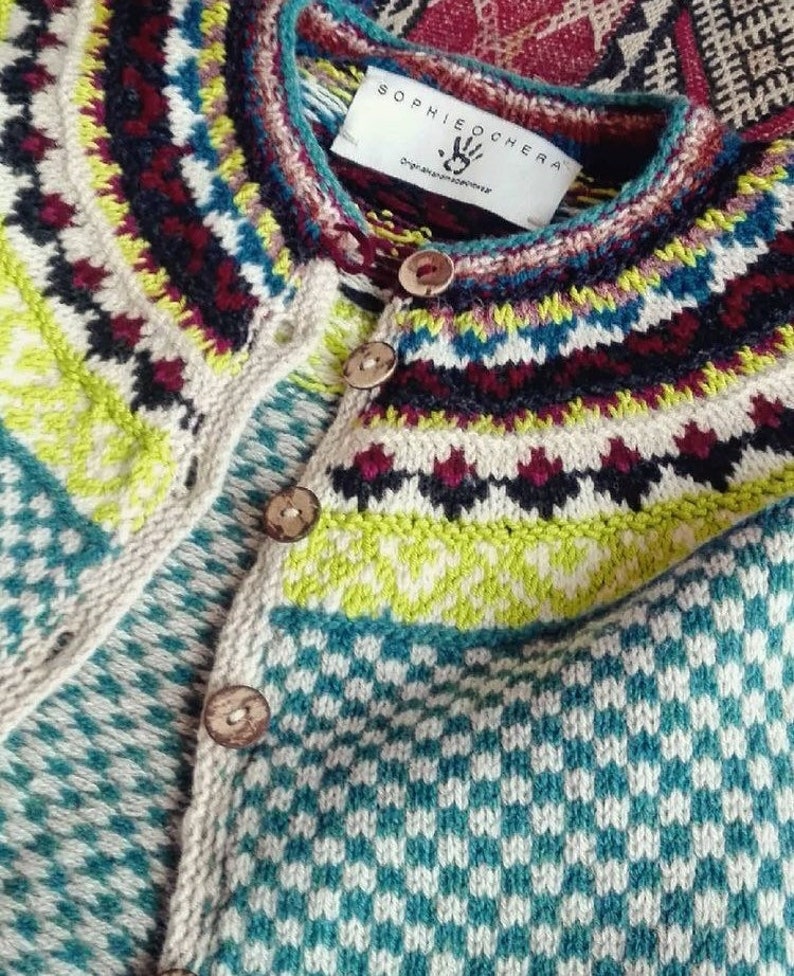 Puede incluir: Un c&aacute;rdigan de punto con un patr&oacute;n geom&eacute;trico colorido y botones de madera. El c&aacute;rdigan tiene una etiqueta blanca con el texto "SOPHIE OCHER A" y un logotipo de huella de mano.