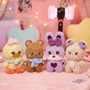 Pode incluir: Quatro brinquedos de pelúcia de crochê feitos à mão: um pato amarelo, um urso marrom, um coelho roxo e um urso bege. Cada um tem um laço e um cachecol únicos. Os brinquedos são dispostos em uma superfície rosa, com um fundo rosa e luzes de fadas.