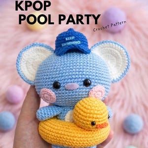 May include: A light blue crocheted koala amigurumi wearing a blue baseball cap with the text KEEP SWIMMING embroidered in white. The koala has pink cheeks, black eyes, and is inside a yellow crocheted duck-shaped pool floatie.
