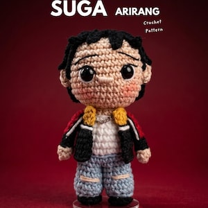 Pode incluir: Boneca amigurumi Suga de crochê, com cabelo preto, jaqueta preta com detalhes em vermelho e amarelo, jeans azul e corrente prateada. A boneca está em um suporte transparente. A imagem inclui o texto "SUGA ARIRANG Crochet Pattern".