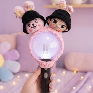 May include: A black and pink light stick with a crochet cover featuring two cartoon characters wearing black hats. The light stick has a glowing orb with the text "BTS" and is held by a person.