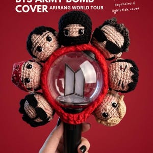 Puede incluir: Una funda de lightstick BTS Army Bomb roja y negra con cabezas de personajes de ganchillo. La funda es parte de un paquete con llaveros. El texto en la imagen dice "BTS ARMY BOMB COVER ARIRANG WORLD TOUR".