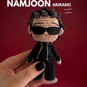 May include: A crocheted amigurumi doll of a person wearing sunglasses and a black outfit. The doll has dark hair and is held in front of a red background. The text "EBOOK NAMJOON ARIRANG Crochet Pattern" is displayed above the doll.