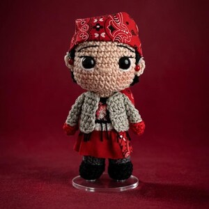 May include: A crocheted doll with large black eyes, wearing a red bandana with a white paisley pattern, a light gray textured cardigan, a red skirt, and black shoes. The doll has small red bead earrings and stands on a clear circular base.