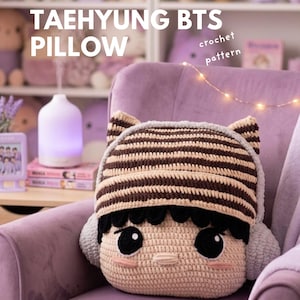 Pode incluir: Uma almofada de crochê feita à mão com um rosto de desenho animado com olhos grandes, um chapéu listrado e protetores de ouvido. A almofada é bege, marrom e preta. O texto "TAEHYUNG BTS PILLOW" é exibido acima da almofada.