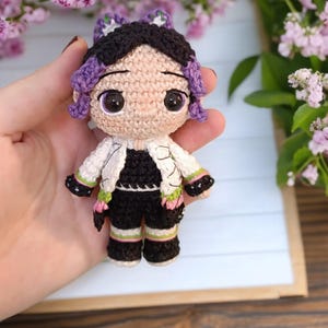 Könnte beinhalten: Häkelpuppe mit schwarzen Haaren, lila und weißer Kleidung und großen braunen Augen. Die Puppe trägt eine Jacke mit weißen und schwarzen Mustern und eine schwarze Hose mit grünen und weißen Streifen.