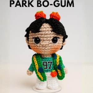 May include: A crocheted amigurumi doll of Park Bo-gum, wearing a green and yellow jacket with the number 97. The doll has black hair, orange hair accessories, and is wearing a green shirt and white pants. The image includes the text "EBOOK PARK BO-GUM".