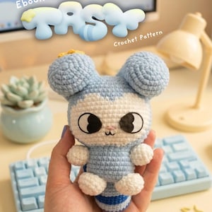 May include: A light blue and white crocheted amigurumi figure with large black eyes, a small embroidered mouth, and rounded ears. The text Ebook TASY Crochet Pattern is visible above the figure, with social media handles and a small logo below.