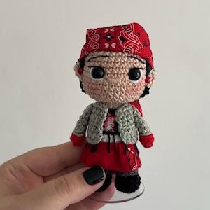 May include: A small crocheted doll featuring a tan face with large black eyes, wearing a red bandana with a white paisley pattern, a light gray textured vest, a red skirt, and black shoes. The doll is standing on a clear circular base.
