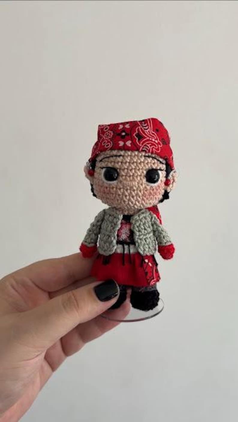 May include: A small crocheted doll featuring a tan face with large black eyes, wearing a red bandana with a white paisley pattern, a light gray textured vest, a red skirt, and black shoes. The doll is standing on a clear circular base.
