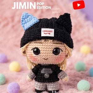 Puede incluir: Una muñeca amigurumi de ganchillo con cabello rubio, un gorro negro con orejas de gato y una camiseta negra con el texto ARIRANG WORLD TOUR. La muñeca tiene grandes ojos negros, mejillas rosadas y una pequeña sonrisa. Texto: EBOOK JIMIN POP EDITION.