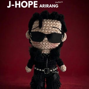 May include: A crocheted amigurumi doll of J-Hope wearing black sunglasses and a black outfit. The doll has a light brown complexion and black hair. The image includes the text "EBOOK J-HOPE ARIRANG Crochet Pattern" and social media handles.