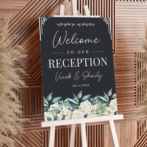 May include: Black welcome sign with white text that reads "Welcome to our Reception Vivek & Shaily 09.14.2024". The sign is decorated with green and white floral illustrations.