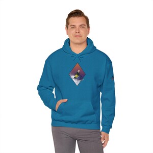 Op de afbeelding: Een turquoise blauwe hoodie met een ruitvormige afbeelding van een persoon die snowboardt van een berg af. De afbeelding heeft een kleurthema van paars, blauw en oranje.