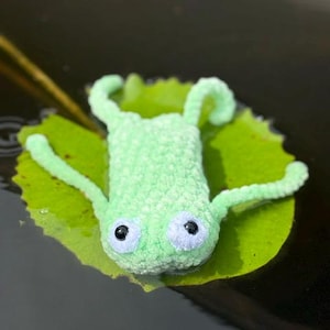 May include: A light green crocheted frog toy with a soft, fuzzy texture. The frog features two large, round white eyes with black pupils and four thin, extended limbs. It is positioned flat on top of a vibrant green lily pad floating in dark water.