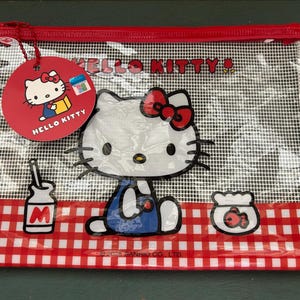 May include: A rectangular clear mesh zipper pouch featuring a red and white gingham pattern at the bottom. The front displays a Hello Kitty graphic, a milk bottle, and a fishbowl. A round red tag with Hello Kitty branding is attached to the zipper.