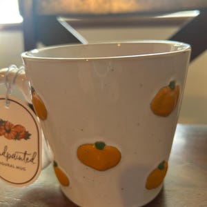 May include: A white ceramic mug with a speckled finish and raised, orange pumpkin motifs. A tag attached to the handle features a graphic of pumpkins and the text "handpainted FIGURAL MUG". The mug has a wide, cylindrical body and a sturdy handle.