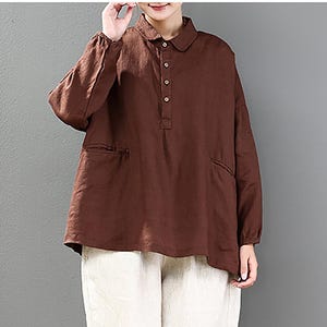 May include: A brown long-sleeved shirt with a collared neckline and button closure, worn with off-white, loose-fitting trousers. The shirt features two front pockets. The outfit is completed with white trainers. The clothing appears to be made of a soft, natural fabric.