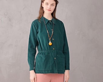 May include: A woman in a dark green corduroy jacket with a button-up front, two front pockets, and a cinched waist. She is wearing pink trousers and brown boots. A long beaded necklace with a yellow pendant is visible.