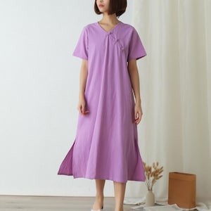 Puede incluir: Un vestido de color lavanda, de largo hasta la rodilla, con cuello en V y mangas cortas. El vestido tiene un cierre de botón en el escote y una abertura lateral a cada lado.