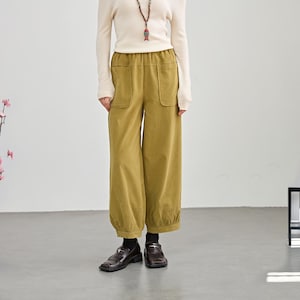 May include: A woman wearing a cream-colored long-sleeve sweater and olive green corduroy pants. The pants have large pockets and a gathered hem. A long beaded necklace with a fish pendant is worn. The outfit is paired with black shoes.