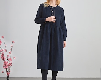 May include: A navy blue corduroy dress with long sleeves and a button-down front. The dress falls to mid-calf length. The person is wearing black leggings and dark shoes. A black vase with pink flowers is on the left.