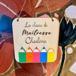 Peut inclure: Panneau en bois avec le texte "La classe de Maitresse Charlène" et des illustrations de crayons colorés. Le panneau est carré avec des coins arrondis et une ficelle pour l'accrocher. Les crayons sont verts, orange, roses, bleus et jaunes.