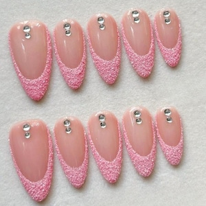 Puede incluir: Conjunto de diez uñas artificiales en tono rosa claro, cada una adornada con una punta de purpurina rosa brillante y una pequeña gema transparente. Las uñas, de forma ovalada, están dispuestas sobre una superficie blanca.