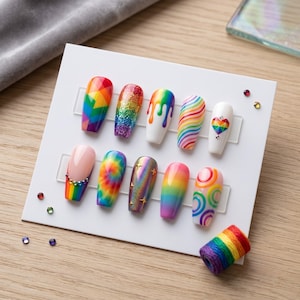 Pode incluir: Uma exibição de dez unhas artificiais com vários designs. As unhas apresentam padrões de arco-íris, glitter, tie-dye e outros designs coloridos. Algumas unhas têm detalhes de coração e estrela. Também está presente uma pequena bobina de linha arco-íris.