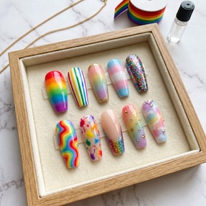 May include: A wooden tray displays ten artificial nails with various designs. The nails feature rainbow gradients, stripes, glitter, and other colorful patterns. Some nails have gold accents and star embellishments. A rainbow ribbon and a bottle are in the background.