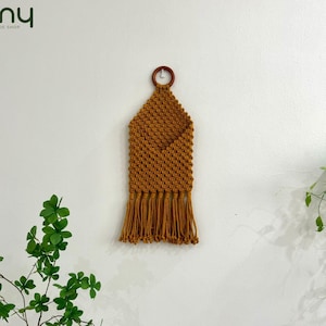 May include: A mustard yellow macrame wall hanging with a pocket and fringe. The hanging is attached to a white wall with a wooden ring.