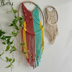 May include: Two macrame wall hangings. One is a butterfly shape with red, yellow, and teal sections, wooden beads, and a wooden hoop. The other is a cream-colored butterfly shape with wooden beads and a wooden hoop.
