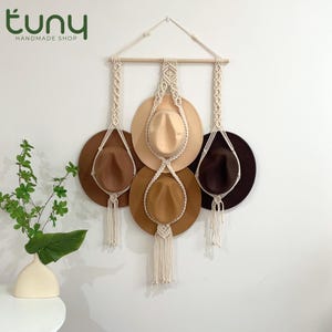 May include: A macramé hat rack displaying four hats in shades of brown and beige. The hats are suspended from a wooden dowel and a triangular macramé structure. The hats are displayed against a white wall.