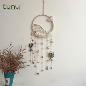 May include: A handmade macrame whale dreamcatcher with a wooden hoop and hanging strands. The whale is crafted with cream-colored macrame, and the design includes colorful accents and decorative elements. The shop name "tuny handmade shop" is in the top left corner.