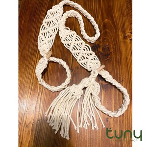 Puede incluir: Una correa de macramé color blanco roto con intrincados patrones de nudos, dos bucles circulares y dos cuentas de madera. La correa termina en un fleco largo y grueso hecho de múltiples cordones de algodón colgantes, creando un diseño texturizado.