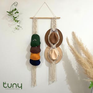 Könnte beinhalten: Makramee-Hutständer mit Holzstab und Aufhängeschnur. Er zeigt eine Reihe von Baseballkappen in Grün, Braun, Orange und Marineblau. Zwei Fedora-Hüte in Braun und Beige sind ebenfalls zu sehen. Die Wand ist weiß.