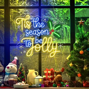 May include: A neon sign with the text "Tis the season to be Jolly" in yellow and blue. The sign is decorated with snowflakes and holly. A small Christmas tree and other holiday decorations are in front of a window.