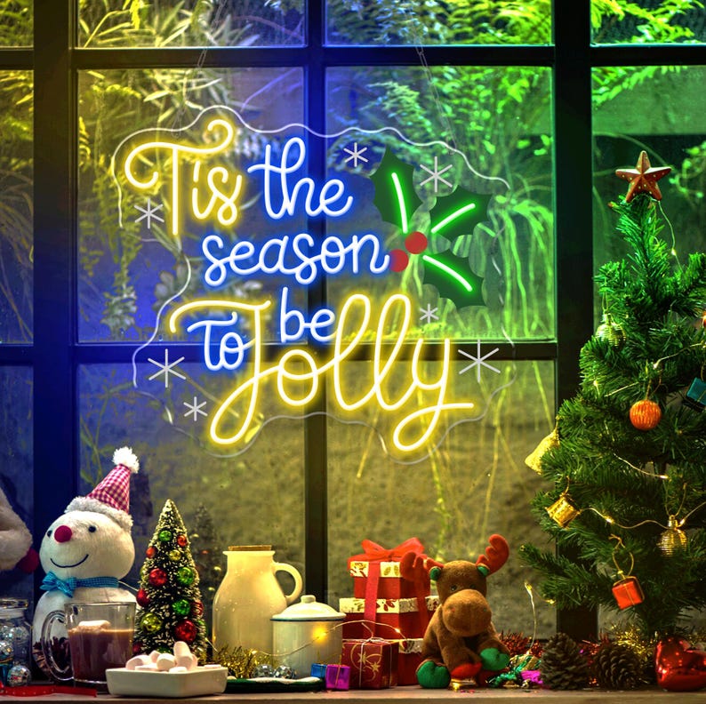 May include: A neon sign with the text "Tis the season to be Jolly" in yellow and blue. The sign is decorated with snowflakes and holly. A small Christmas tree and other holiday decorations are in front of a window.