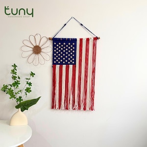 Puede incluir: Un tapiz de pared de macramé hecho a mano con la bandera estadounidense. La bandera presenta un campo azul con estrellas blancas y rayas rojas y blancas. Se suspende de un dowel de madera con un cordón azul.