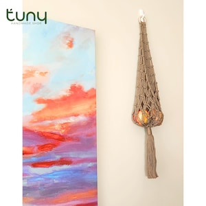 May include: A handmade macrame fruit hammock in a light brown color, hanging on a white wall. The hammock is filled with oranges. A colorful abstract painting is on the left. The shop logo "tuny handmade shop" is in the top left corner.